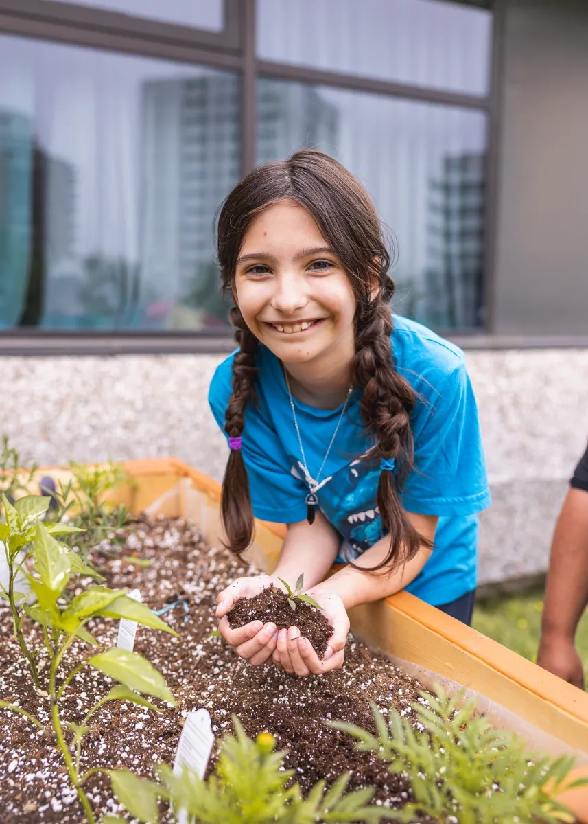 A young girl is holding a handful of dirt with both hands and smiling at the camera. She is bent over a raised garden bed with greenery sprouting.
