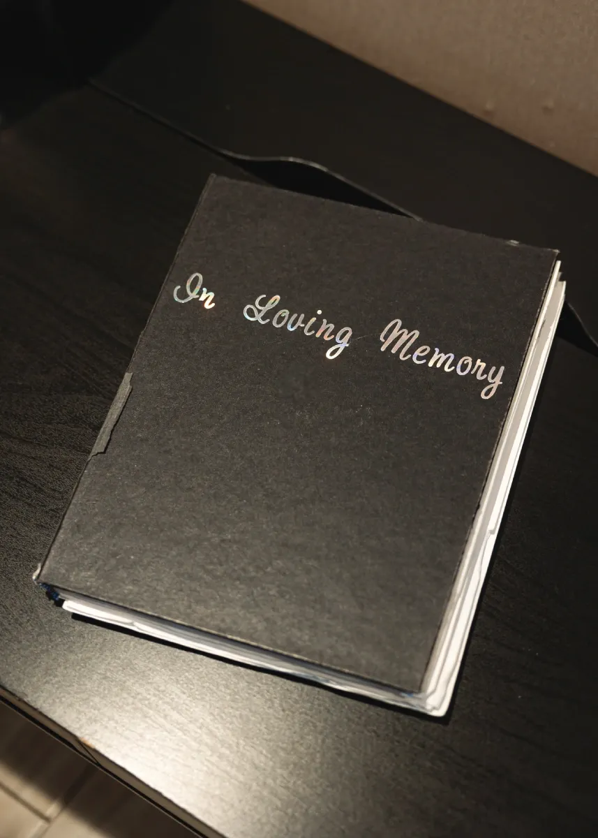 A black binder sits on a dark brown table. The front of the binder reads "In Loving Memory" in cursive.