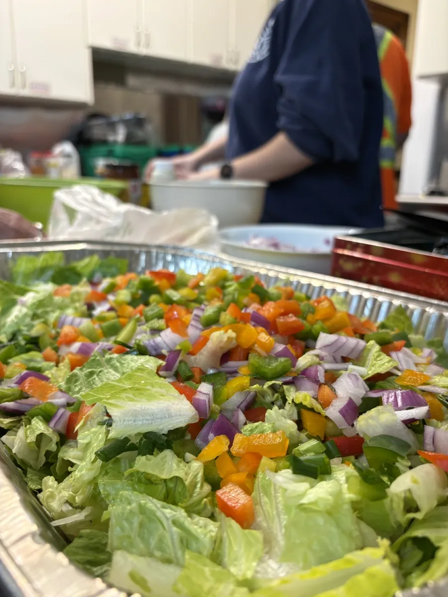 Close up image of a salad. In the background you can see a person preparing more food in a bowl.