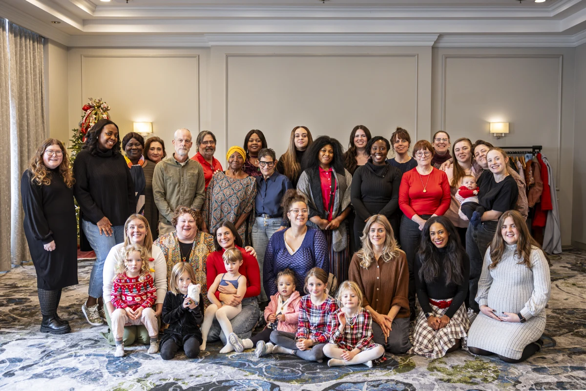 A large group of people are posing in a hotel ballroom. There are people kneeling, children sitting on the floor and a large group standing behind them. A Christmas tree can be seen in the background.