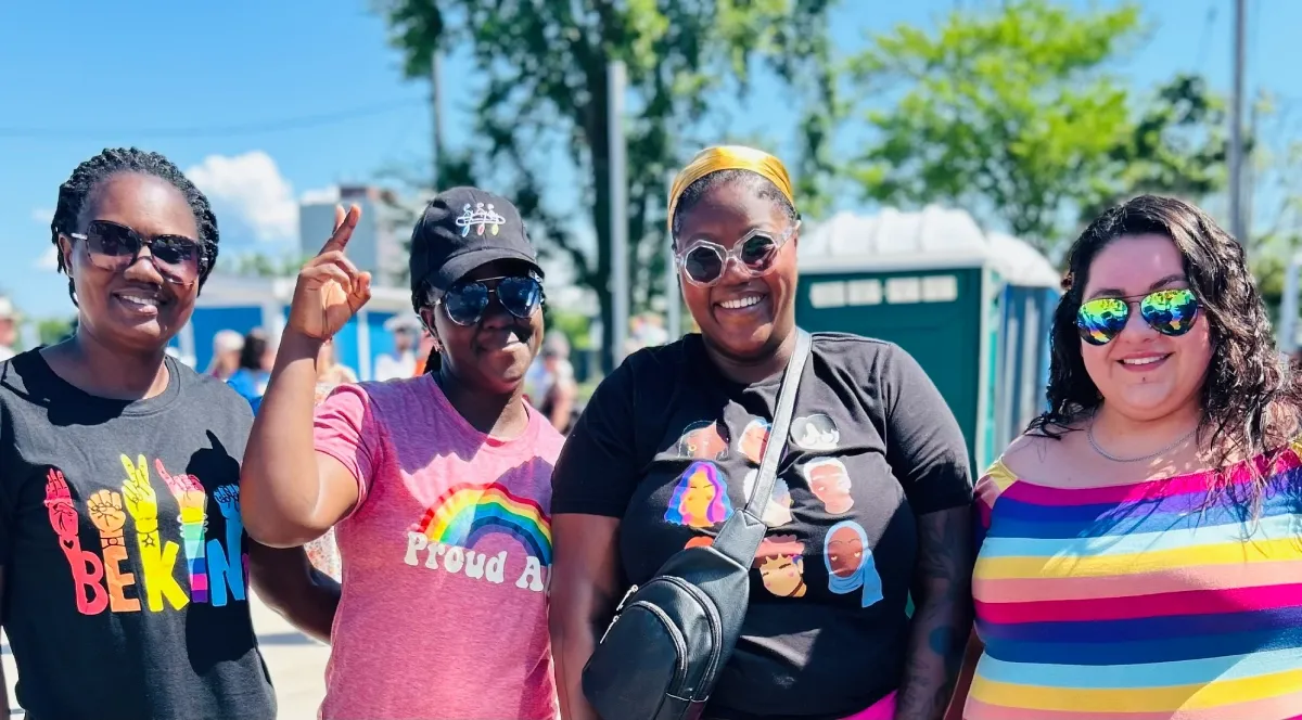 Four people stand outside in the sunshine posing for a photo. They are wearing various Pride gear.