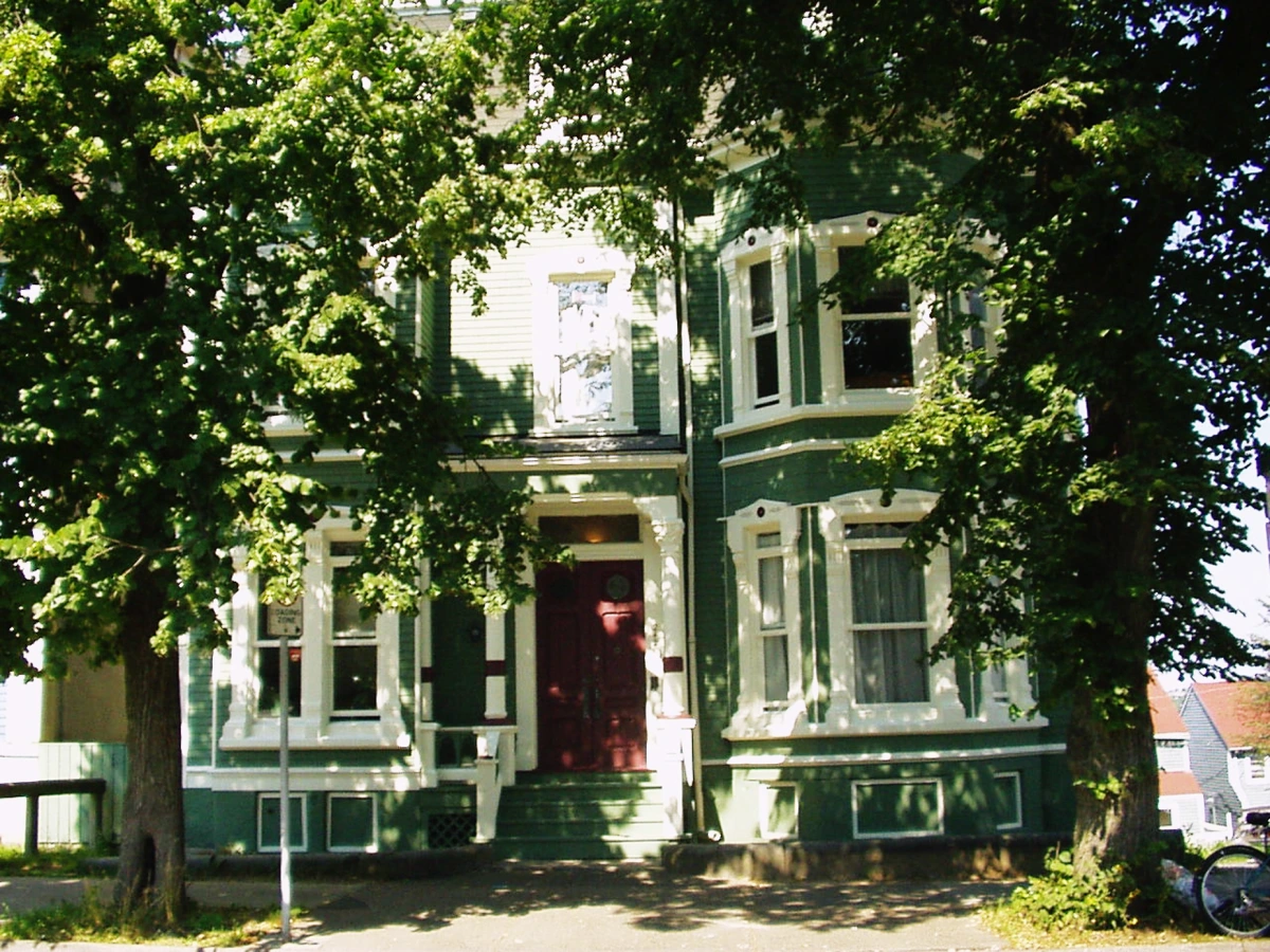 Image of a green siding house with a red door in the sunlight. Trees frame the house on either side.