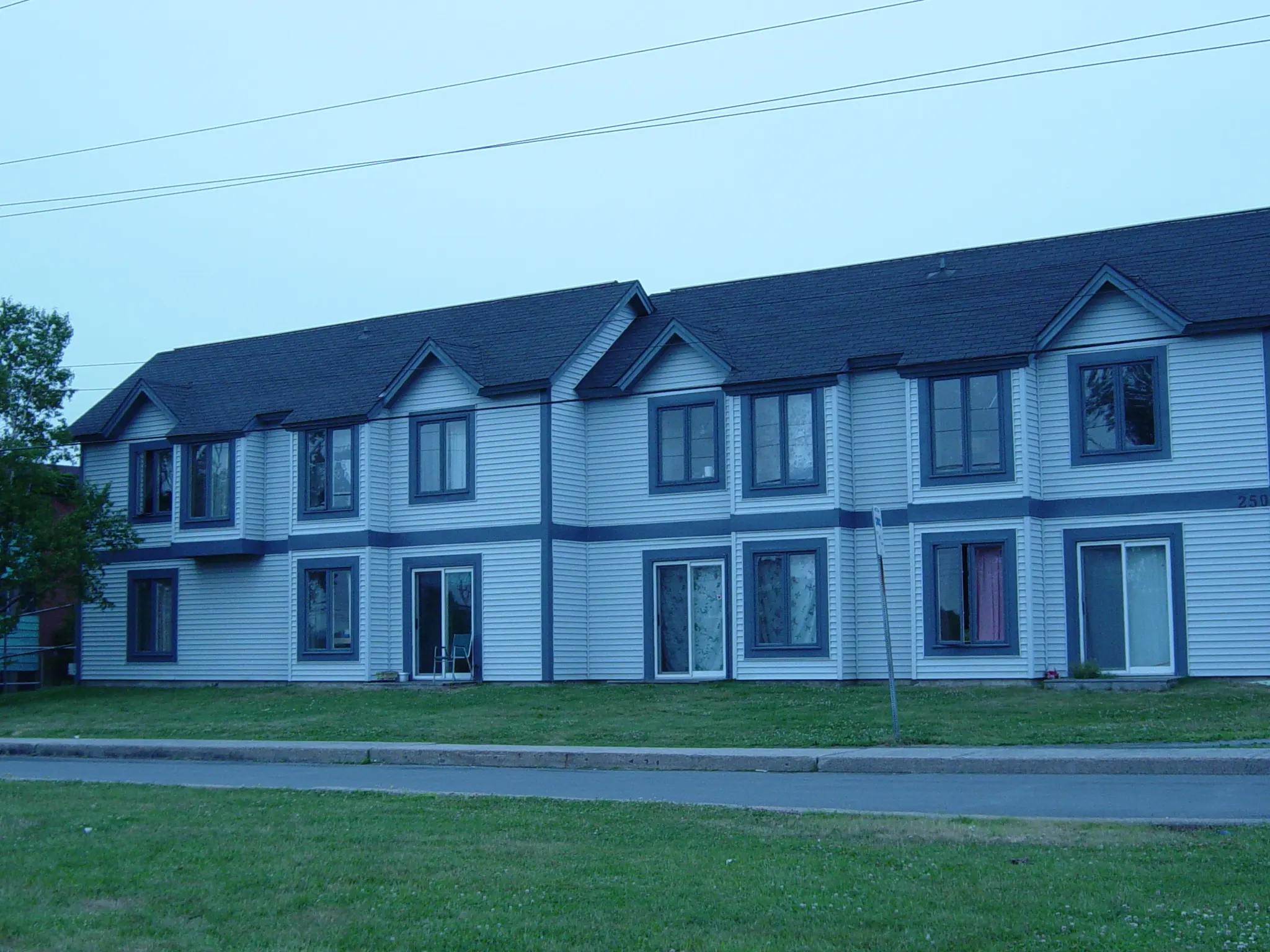 Image of a multi-unit residential building with white siding. A grass lawn grows in front of the building and a road can be seen in the foreground. 
