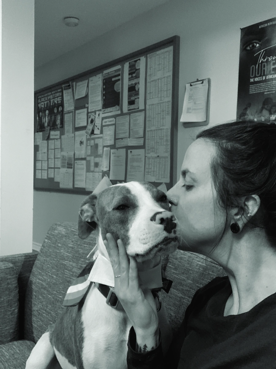 A black and white photo of a women kissing a dog on the cheek. The background has a long notice board with many notices attached. 