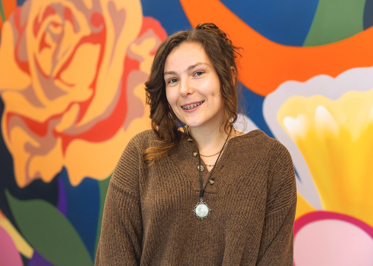 A smiling person with long hair that is clipped up on the back poses in front of a brightly colour flower mural wall. 