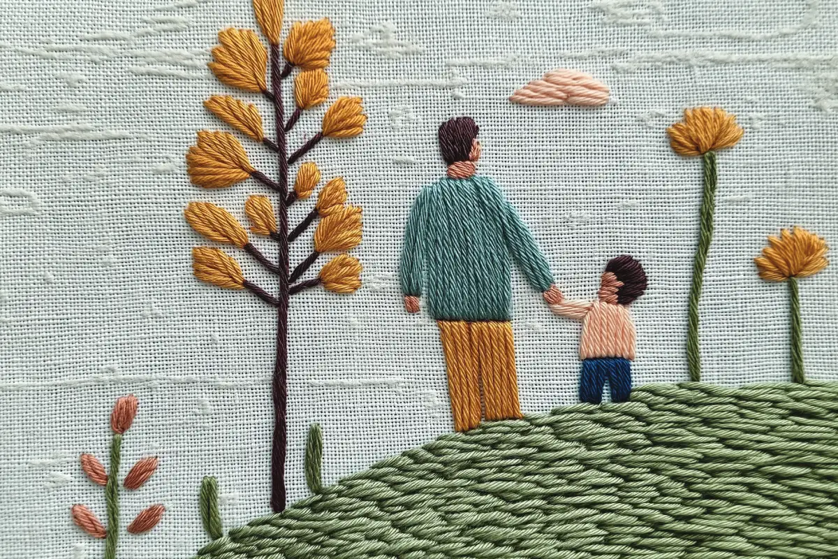 Embroidered image of a parent and a child holding hands in a field. There are a number of yellow and pink flowers and a large yellow tree. 