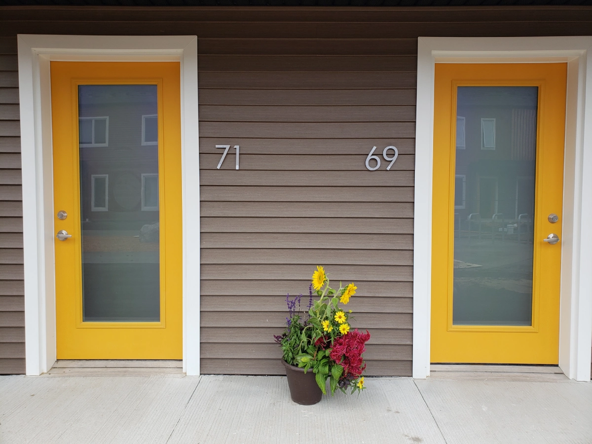 A brown siding wall with two yellow doors. The door on the left has the address "71", and the door on the right has the address "69". There is a potted plant with yellow, red, and purple flowers on the ground in between the doors.