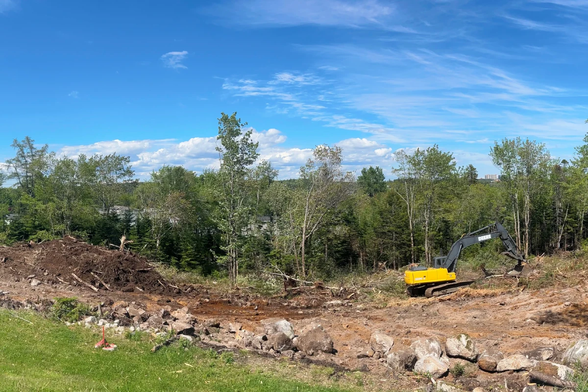 A bright yellow digger is pictured clearing rocks and dirt. The site is surrounded by trees and a bright blue sky.