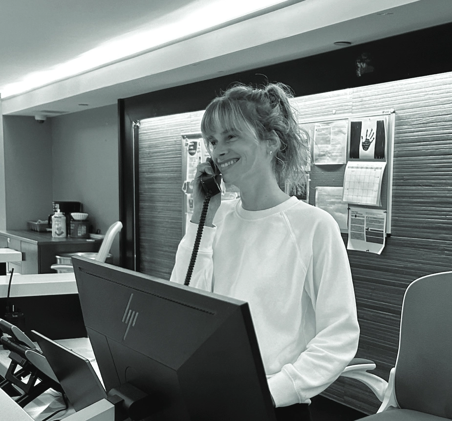 A black and white photos of a person with long hair tied back in a ponytail is smiling and holding a corded phone. Computer monitors can be seen in the foreground and the background has a notice board.