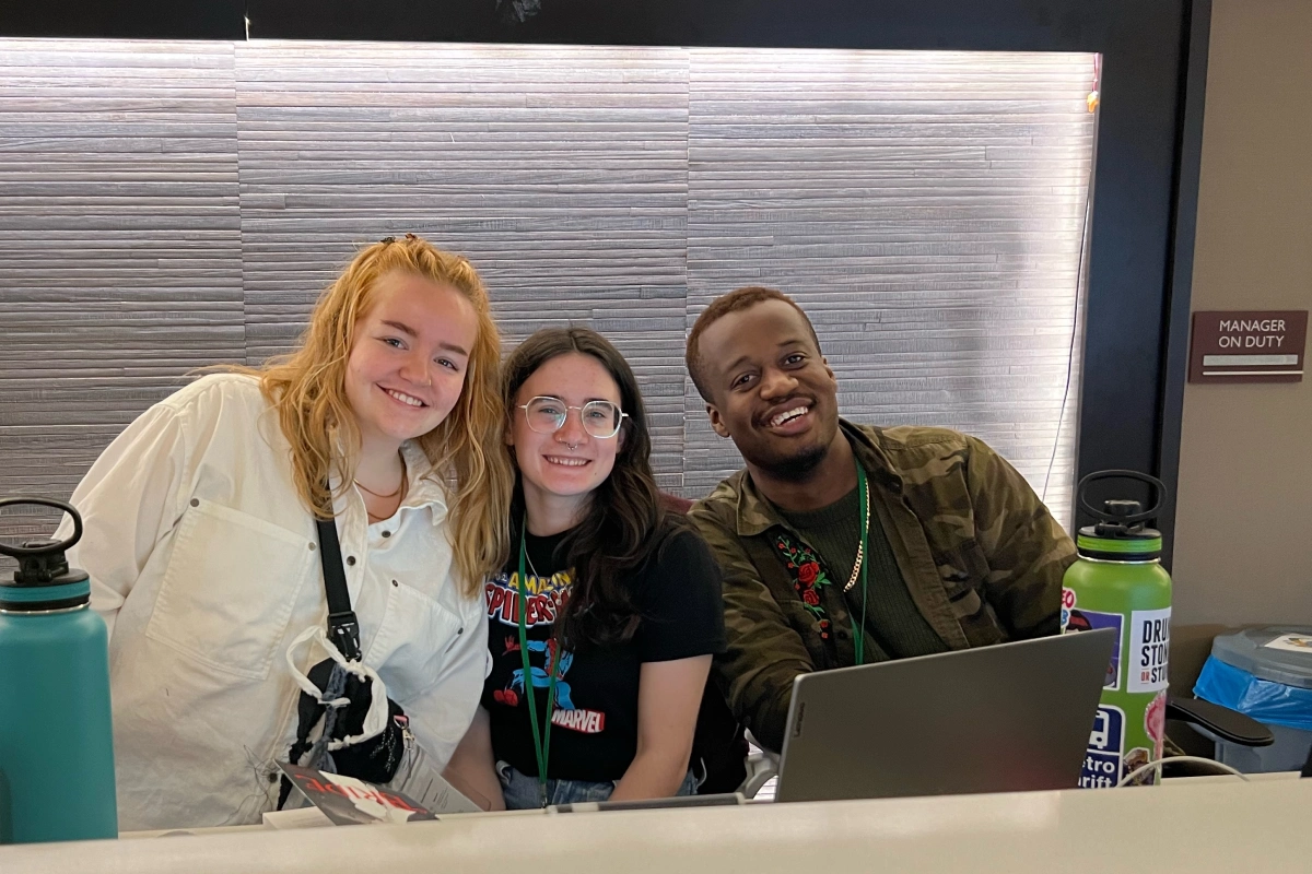 Three people stand behind a reception desk smiling at the camera. Water bottles can be seen on the desk.