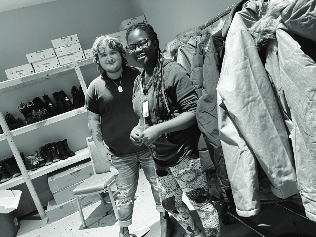 A black and white photo of two people standing in a storage room. Shoes can be seen on shelves in the back of the room and a rack of coats in to the right.