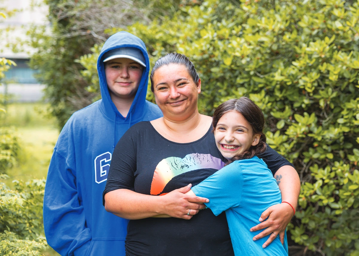 A mom and her two kids pose hugging each other and smiling at the camera. They are pictured outside with green bushes in the background.