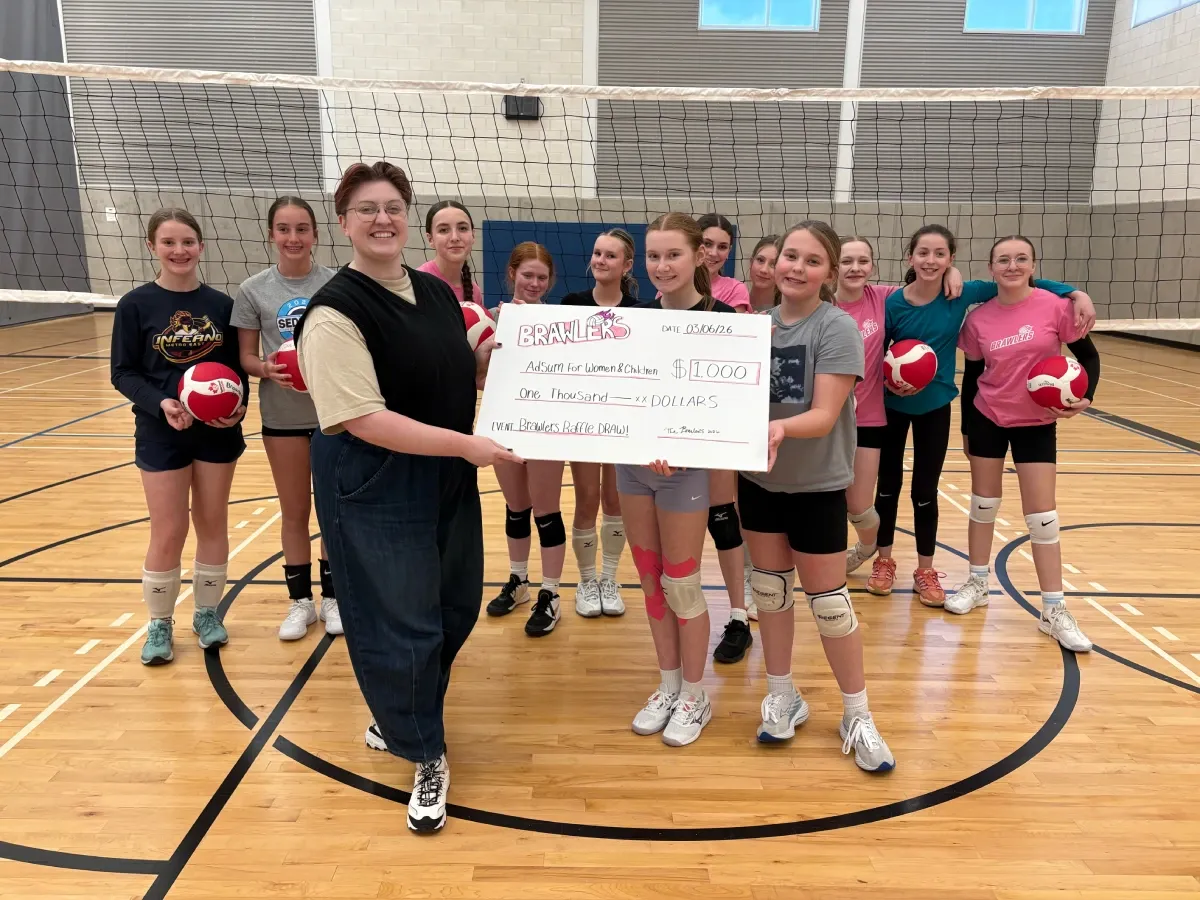 A group of people pose for a photo in front of a volleyball net. 10 young girls pose with volleyballs in the background and in the foreground two young girls and an adult hold a cheque for $1000 for Adsum.