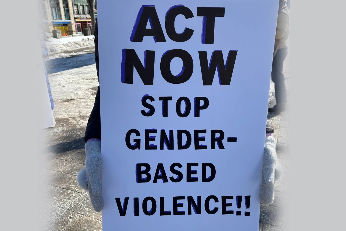 A protest sign reading "Act Now Stop Gender-Based Violence!!" is held in the centre of the photo. The sign holder is wearing gloves and standing outside on a sidewalk in the winter.