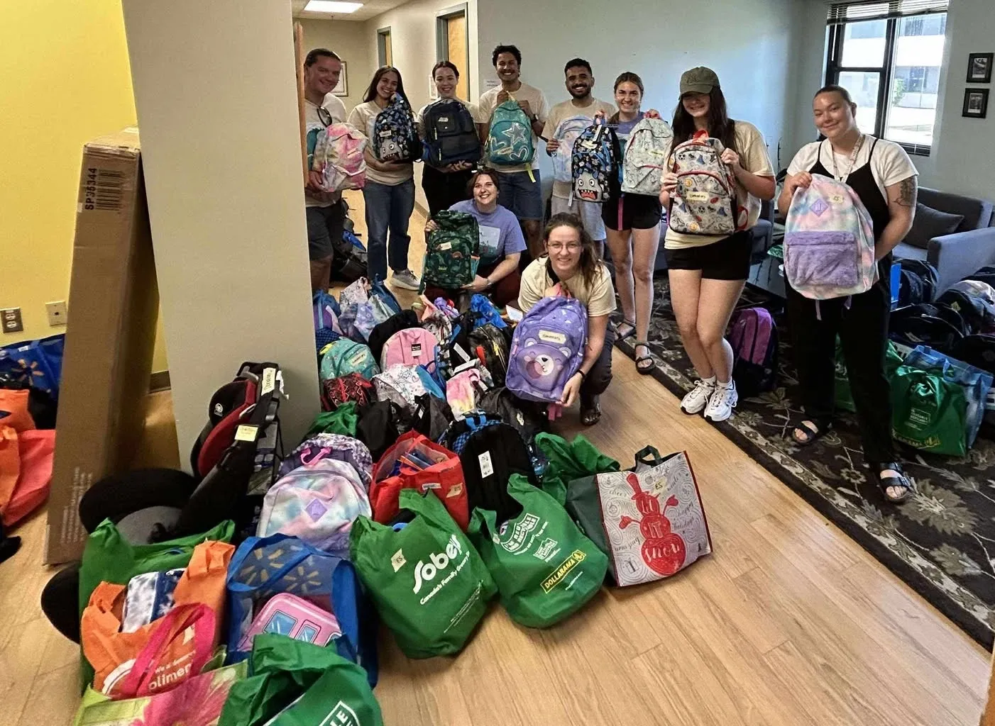10 people are posing holding backbacks and smiling at the camera. On the floor in front of them are more backpack and other bags of donations.