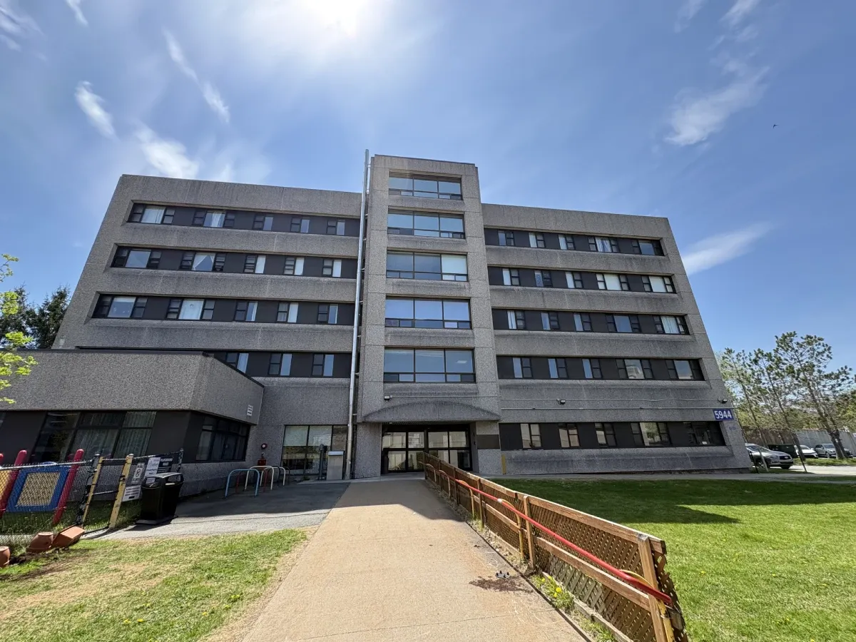 A paved pathway stretches in front of the camera with a railing on the right side leading to a six-story apartment building in the center of the image. Blue sky surrounds the building and playground gates lead off to the left side of the image. 