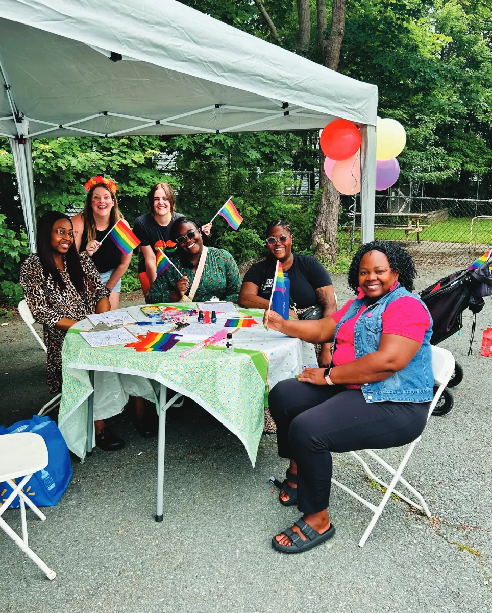 Six people stand and sit around a table under a tent outside. They are smiling at the camera and waving varioud small pride flags. The table holds more pride flags.