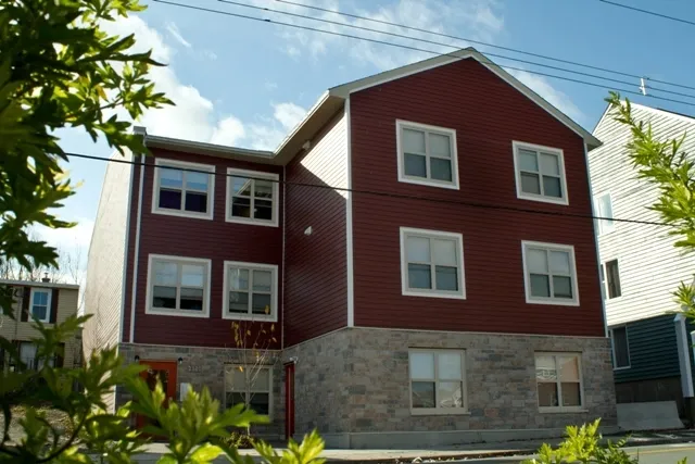 A three-story building is framed by greenery. The top floors are red siding and the bottom floor is grey brick.