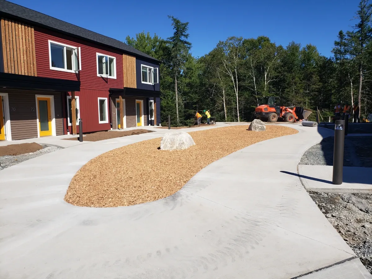 A modern multi-unit residential building faces a curving pathway and garden bed. In the background, workers and a tractor stand near a line of trees.
