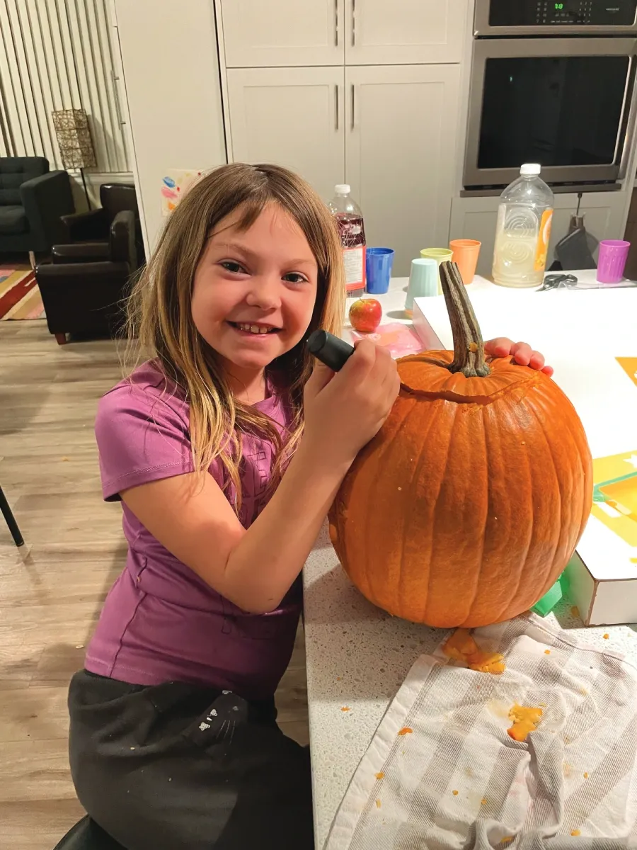 A young girl is pictured carving a pumpkin. She is smiling at the camera.