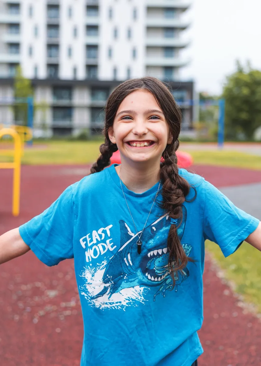 A young girl wearing blue is smiling at the camera and reaching her arms out to the side. A playground can be seen in the background.