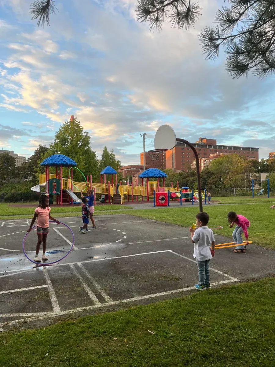 Five children are pictured playing on an outdoor basketball court. Three of the children are playing with brightly coloured hula-hoops and one child is drinking a juicebox. A playground is pictured in the background.